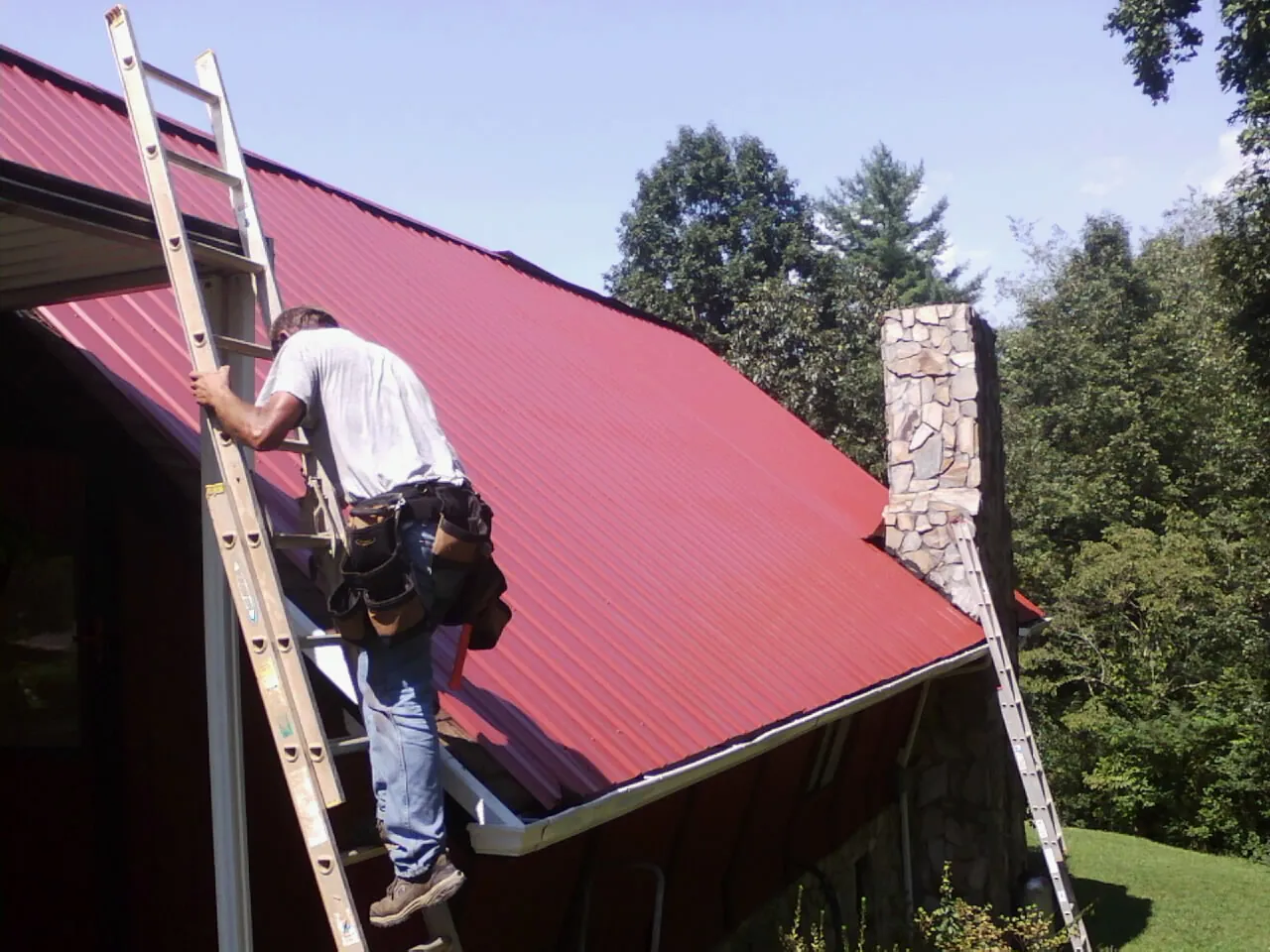 Roofer on a ladder beside a red metal roof and stone chimney — Soffit & Fascia Repair by Roofing Brothers in Vonore